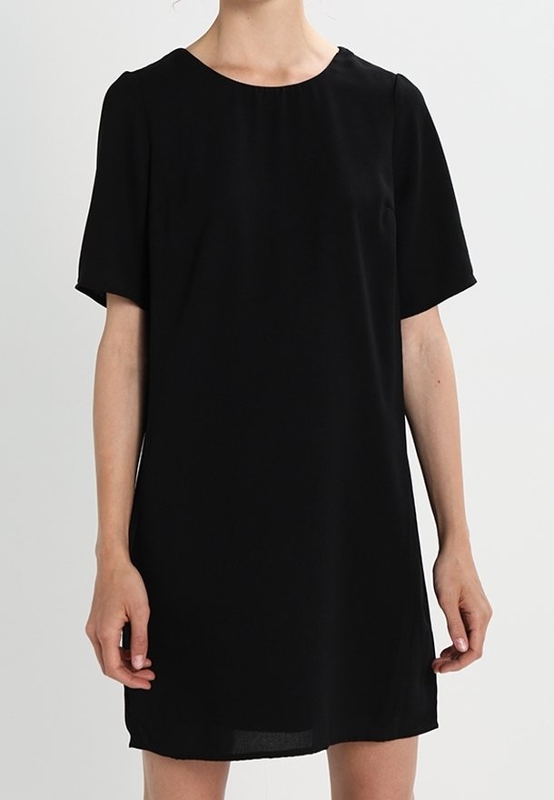 Femme portant une robe noire unie à manches courtes avec un col rond sur un fond neutre.