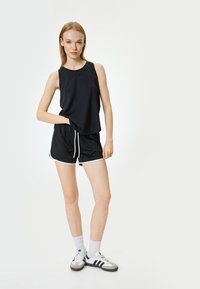Top negro sin mangas combinado con pantalones cortos negros que tienen cordones y detalles en blanco. El modelo lleva calcetas blancas y zapatillas de deporte en blanco y negro.