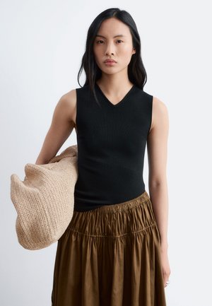 Jeune femme aux longs cheveux noirs portant un haut noir sans manches et une jupe marron, tenant un grand sac tissé beige sur un fond uni.