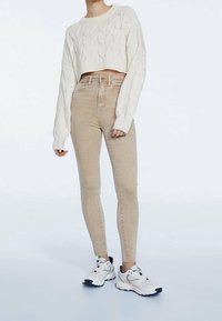 Suéter corto de punto trenzado color crema con mangas largas, combinado con jeans skinny beiges de tiro alto y zapatillas blancas con detalles en negro.
