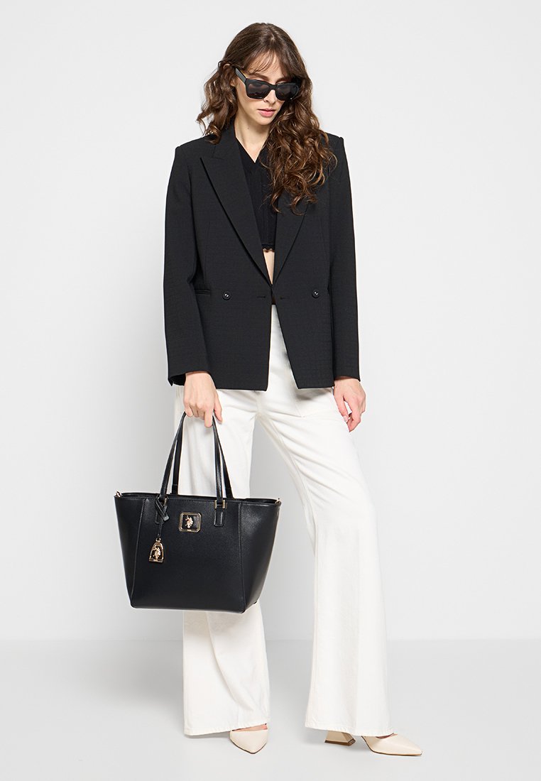 Bolso tote negro con forma estructurada, cuero suave y herrajes dorados. Presenta dos asas superiores y un charm decorativo.