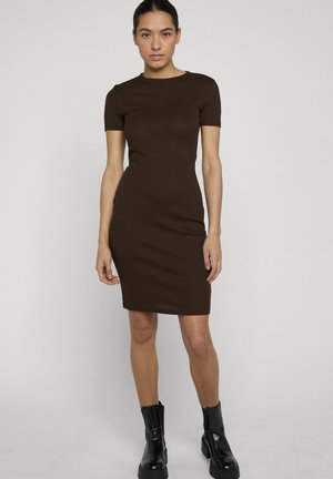 Nő áll, sötétbarna, testhez simuló, rövid ujjú, térdig érő ruhát visel, valamint fekete középmagasságú csizmát, egyszínű világos háttér előtt.