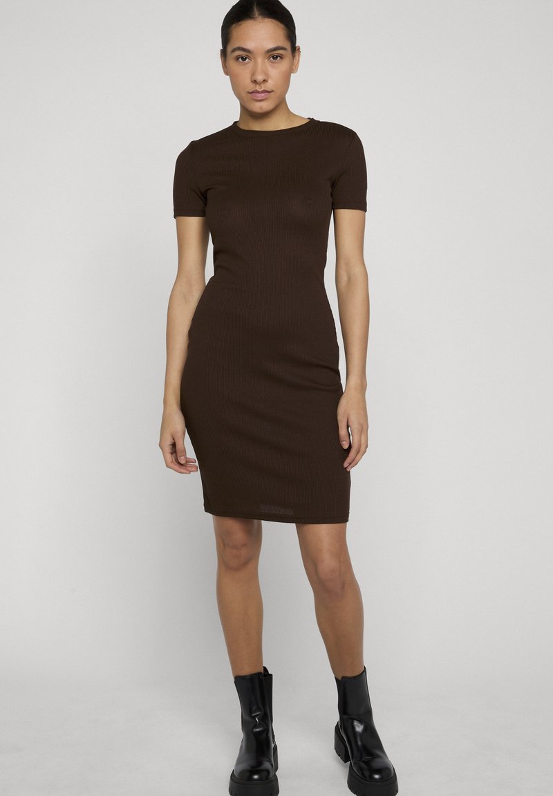 Femme debout portant une robe moulante à manches courtes, marron foncé, longueur genou, et des bottes noires mi-mollet, sur un fond clair uni.