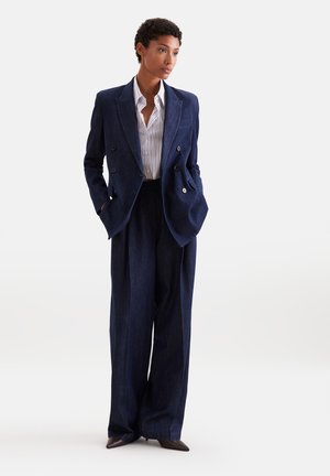 Femme debout, les mains dans les poches, portant un blazer croisé bleu marine, un pantalon large assorti, une chemise rayée et des chaussures pointues foncées.