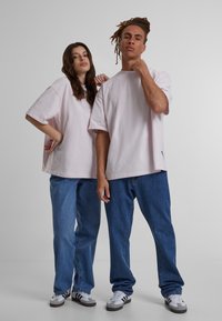 Rožinės per didelės t-shirts, laisvi mėlyni džinsai ir balti sportbačiai su pilkais akcentais. Paprastas dizainas, atsitiktinis siluetas ir švelni audinio tekstūra.