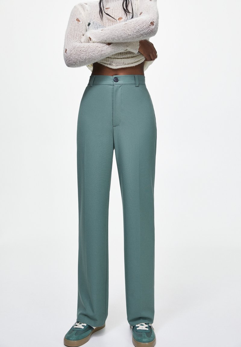 Pantaloni a gamba larga color teal chiaro con una texture liscia, vita alta e chiusura con un singolo bottone, abbinati a sneakers verdi e bianche.