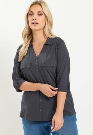 Mujer rubia con una camisa gris oscuro abotonada de mangas tres cuartos enrolladas y pantalones vaqueros azules, de pie con una mano cerca de la cintura.