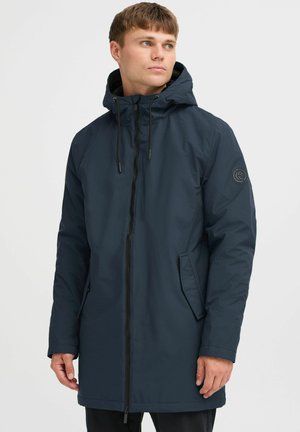 Chaqueta de lluvia larga de color azul oscuro con cierre de cremallera, capucha ajustable y bolsillos laterales. Presenta un parche con el logo en la manga izquierda.