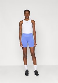 Λευκή αμάνικη μπλούζα, ανοιχτό μπλέ σορτσάκι Nike Pro με λογότυπο στη μέση. Το μοντέλο φοράει μαύρα παπούτσια και λευκές κάλτσες, στέκεται μπροστά σε ουδέτερο φόντο.