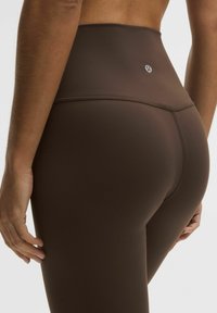 Leggings marrones de talle alto hechos de tela suave y elástica, con costuras mínimas y un logo en la parte trasera.