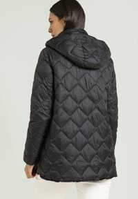 Donna che indossa una giacca nera trapuntata con cappuccio, vista da dietro, abbinata a pantaloni bianchi su uno sfondo semplice.