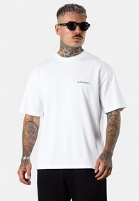 Weißes Baumwoll-T-Shirt mit Rundhalsausschnitt und kurzen Ärmeln, ausgestattet mit einem kleinen schwarzen Logo auf der linken Brust. Glatter Stoff, lockere Passform.