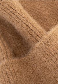 Béžová pletenina s měkkou, načechranou texturou a jemnými žebrovanými vzory. Materiál působí teple a silně, poskytující útulný vzhled.