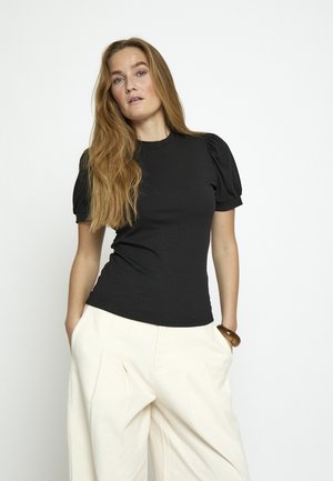 Kobieta o długich włosach w czarnej bluzce z bufkami i szerokich kremowych spodniach, stojąca z rękami w kieszeniach na jednolitym tle.