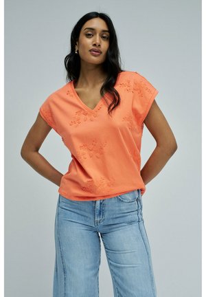 Mujer con cabello largo y oscuro que lleva una blusa coral bordada con escote en V y vaqueros azul claro, de pie con las manos en las caderas.