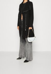 Svart ullblandad kappa med silverfärgade dragkedjor, långa ärmar och sjalkrage; matchad med vida grå jeans och en liten blå mönstrad handväska.