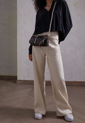 Sort skjorte med knapper, crème brede bukser, hvide sneakers og en lille sort taske med guldaccenter, sat mod en neutral baggrund.