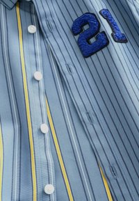 Chemise rayée bleue et jaune avec des boutons blancs et un écusson bleu texturé avec le numéro 21 près de la zone du col.