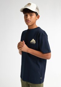 Námořnické modré tričko s krátkým rukávem, s zlatým logem na hrudi. Tkanina vypadá měkce a hladce. Model má na sobě olivově zelené kalhoty.