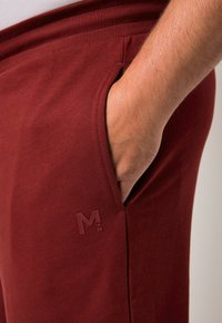 Bordeaux joggings met een gladde textuur, elastische tailleband, zijzak en een klein logo "M+" nabij de zak.