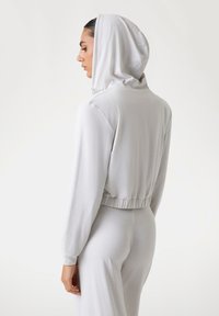 Svijetlo siva hoodie s mekanom teksturom, skraćenim dizajnom, dugim rukavima i kapuljačom na vezanje. Ima elastičnu obrubu na struku i manžetama.