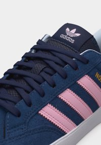 Κοντινό πλάνο ενός αθλητικού παπουτσιού Adidas σε σκούρο μπλε χρώμα, με ροζ ρίγες, σκούρα μπλε κορδόνια και λευκή σόλα.