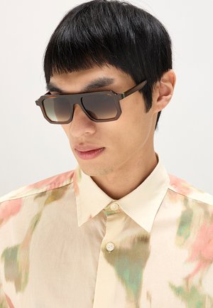 Uomo con capelli lisci e neri che indossa grandi occhiali da sole quadrati in tartaruga e una camicia color crema con motivi verdi e rosa.