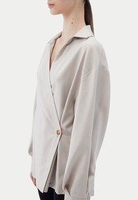 Beige knäppblus med V-ringning, långa ärmar och avslappnad passform; har en enda träknapp och slät textur.