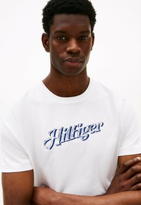 Hvid bomulds t-shirt med blåt broderet logo, klassisk rund halsudskæring, korte ærmer og en afslappet pasform. Glat tekstur, minimalistisk design.