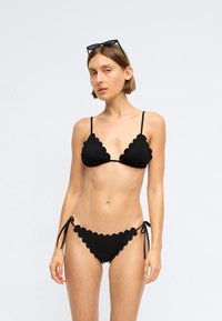 Black bikini set featuring scalloped edges, thin adjustable straps, and side ties on the bottoms. Smooth texture, minimal design.