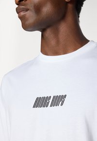 Bílý bavlněný tričko s černým nápisem "Riding Hoops" v nakloněném, moderním písmu, kulatým výstřihem a hladkou texturou.