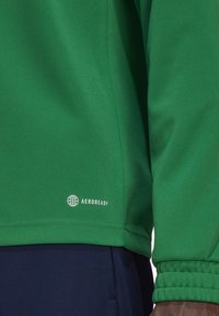 Grünes Sportoberteil aus strukturiertem Stoff. Verfügt über enge Bündchen, einen geraden Saum und ein "AEROREADY"-Logo nahe des unteren Randes.