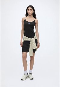 Γυναίκα με μαύρο φόρεμα με λεπτές τιράντες, λευκά αθλητικά παπούτσια, λευκές κάλτσες και ένα ανοιχτόχρωμο ριγέ πουκάμισο δεμένο γύρω από τη μέση, στέκεται μπροστά σε λευκό φόντο.