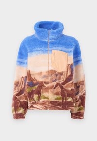 Veste en polaire avec un haut bleu ciel, une poche zippée beige sur la poitrine et un paysage désertique marron présentant des silhouettes de chevaux et de formations rocheuses.