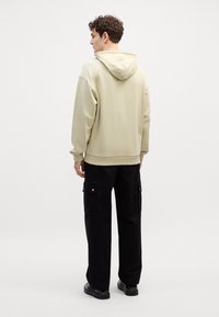 Felpa beige con cappuccio, dalla vestibilità rilassata, con polsini e orlo a costine. Indossata con pantaloni cargo neri dotati di tasche laterali e dettagli con bottoni.