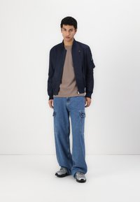Marine bomberjakke, lett stoff, glidelås lukking, brystlomme. Beige crew-neck skjorte, lyse denim cargo bukser, grå sneakers.