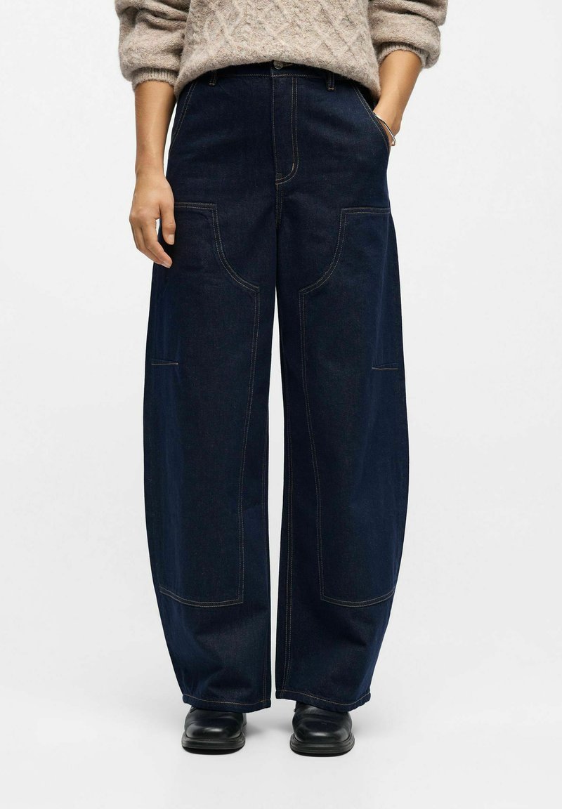 Person, der dunkelblaue weite Jeans mit großen vorderen Aufsatztaschen, einen beigen Pullover und schwarze Lederschuhe trägt, mit einer Hand in der Tasche steht.