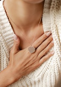 Złoty różowy naszyjnik w kształcie zawieszki z geometrycznym wzorem, zawieszony na delikatnym łańcuszku, wyeksponowany na tle teksturowanego białego swetra.