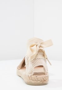 Sandalo espadrilles intrecciato beige con laccio posteriore, tacco aperto e suola in paglia testurizzata. L'accento in nastro di una tonalità più chiara completi il design.