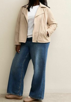 Person iført en beige lynlåstrøje, hvid skjorte, vidtbenede blå jeans og beige sko, stående mod en lys væg.