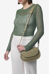 Borsa a tracolla in pelle verde oliva con dettaglio a treccia sul bordo, texture liscia e tracolla regolabile. Portata sopra un top in maglia e pantaloni bianchi.
