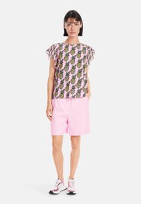 Kurzärmliges Oberteil mit einem rosa Hintergrund und einem wiederholten Ananas-Muster, kombiniert mit hellrosa Shorts und reflektierenden Sneakern.