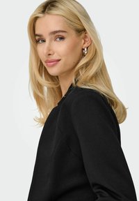 Femme blonde aux longs cheveux portant des boucles d'oreilles argentées en forme d'anneau et un blazer noir, souriant légèrement devant un fond clair uni.