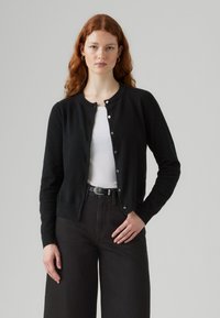 Zwarte cardigan van zacht materiaal, met een ronde hals, knoopsluitingen en lange mouwen, gecombineerd met een witte top en zwarte broek.