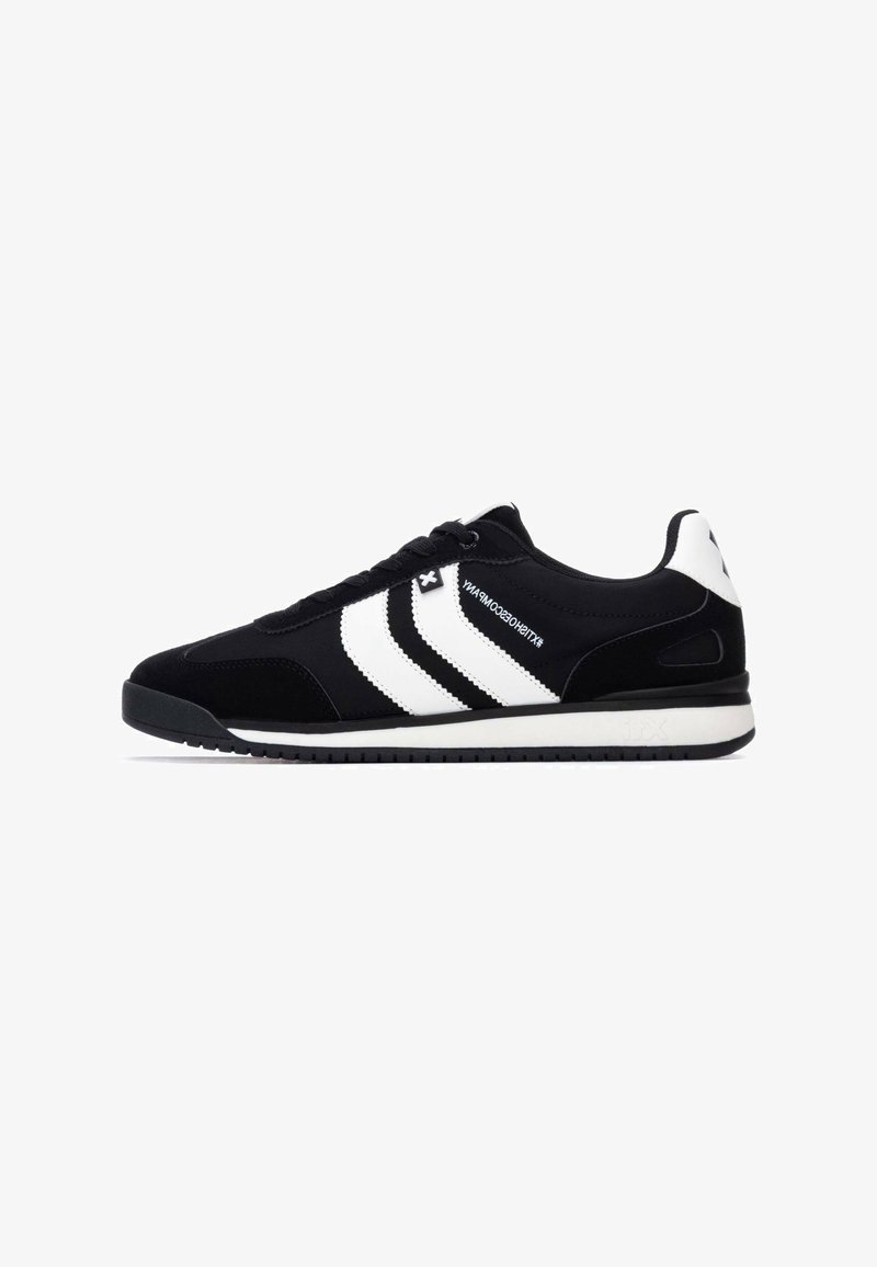 Zapatilla negra con rayas blancas, hecha de ante y malla. Presenta un diseño de cordones y una suela de goma con una superficie texturizada en el talón.