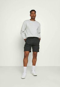 Ung man som står med händerna i fickorna, iklädd en ljusgrå sweatshirt, mörka rutiga shorts, vita strumpor och vita sneakers mot en neutral bakgrund.