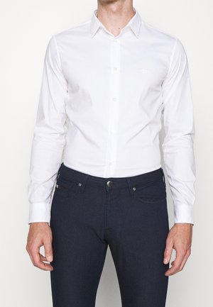 Hombre con una camisa blanca ajustada abotonada, metida dentro de pantalones azul marino oscuro, de pie frente a un fondo liso.