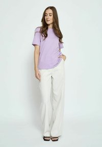 Levandų trumpomis rankovėmis marškinėliai derinami su kreminiais plačiais kelnėmis; modelis stovi su viena ranka kišenėje, demonstruodamas atsipalaidavusį siluetą ir minkštą tekstūrą.