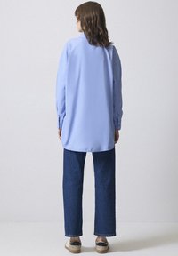 Cămașă albastră deschis cu mânecă lungă, guler, croi drept și mâneci cu nasturi, purtată cu pantaloni din denim închiși la culoare și pantofi casual.