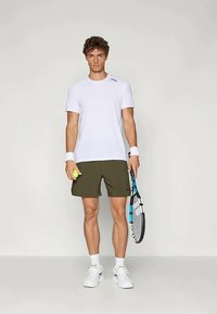 Λευκό αθλητικό T-shirt, ελαιόχρωμο σορτς, λευκά αθλητικά παπούτσια, βραχιόλια, κρατάει μια μπάλα τένις και ρακέτα με μπλε λεπτομέρειες.